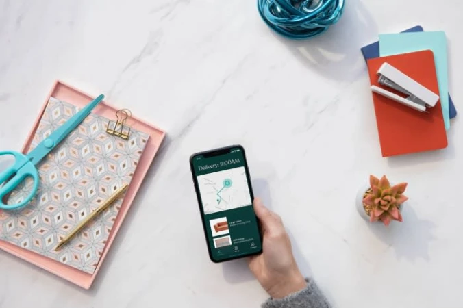 Overhead shot of hand holding smartphone with delivery app, surrounded by scissors, pen, notebooks, stapler, succulent, and yarn on marble.