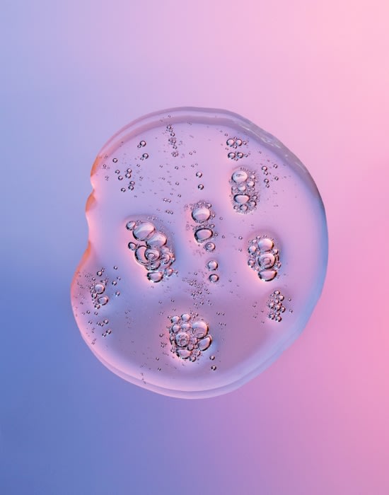 Close-up of a clear, viscous fluid drop with entrapped air bubbles against a gradient background transitioning from blue to pink.