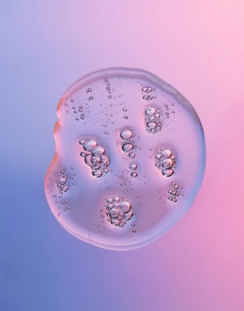 Close-up of a clear, viscous fluid drop with entrapped air bubbles against a gradient background transitioning from blue to pink.