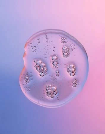 Close-up of a clear, viscous fluid drop with entrapped air bubbles against a gradient background transitioning from blue to pink.