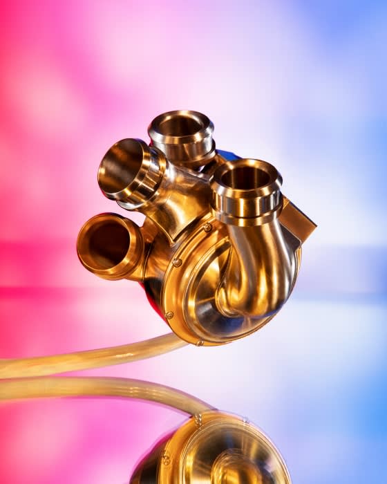 Medical illustration of an artificial heart, a mechanical device with visible pipes, rendered with gold material and a blurred background.