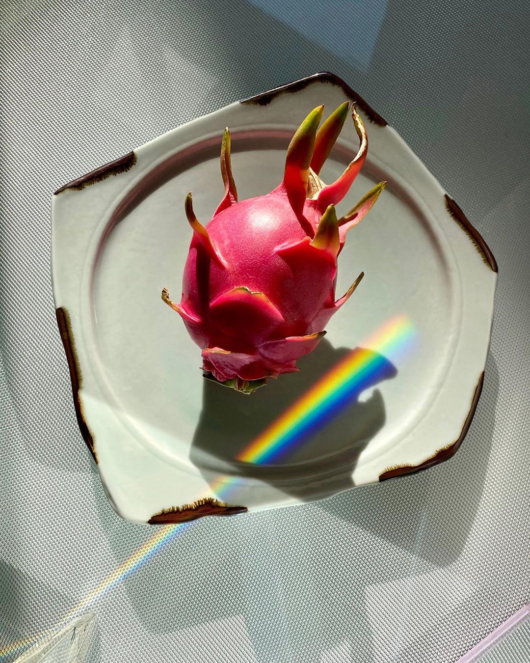 A fresh dragon fruit sits on a white plate with a brown rim, illuminated by a rainbow prism of light, creating a vibrant food still life.