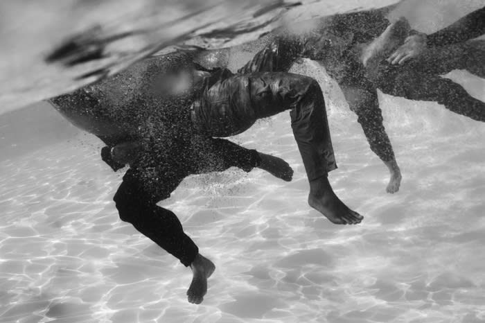 Monochrome underwater photograph of two adults in dark clothing submerged in water with light creating ripple patterns on the sandy bottom.