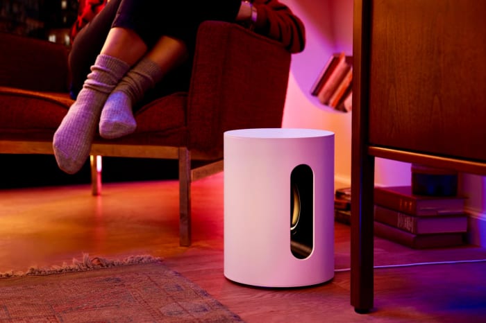 Interior with a white cylindrical speaker, a maroon chair with a person wearing socks, and warm lighting, creating a cozy scene.