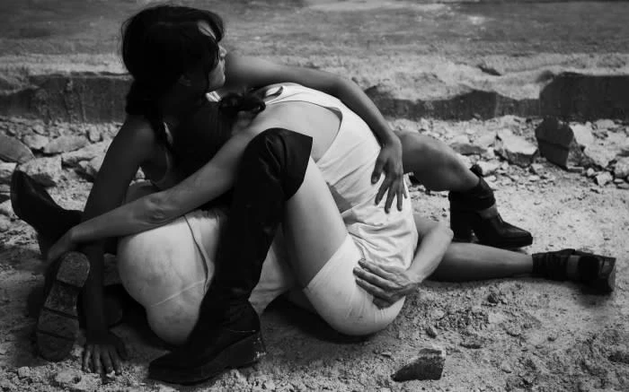 Black and white photo of two women embracing while sitting on rocky ground, one wearing a dress and the other in a long boot.