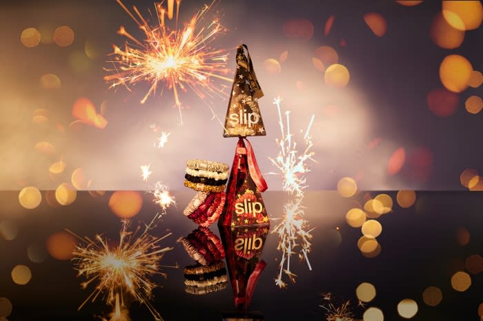 Stacked 'Slip' brand hair accessories and packaging are reflected on a surface amid bokeh and sparkler light effects.