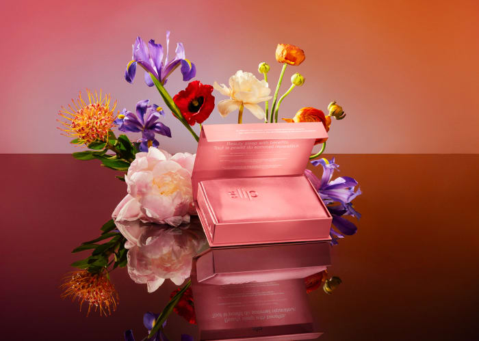 Still life with pink product box surrounded by purple and orange flowers. The composition is set against a pink to orange gradient.