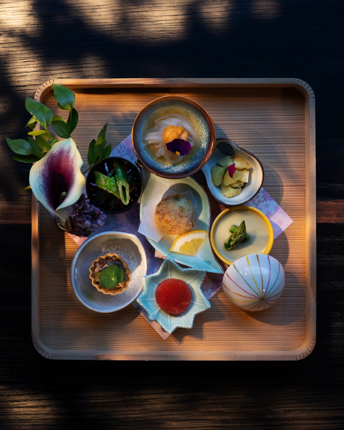 Overhead shot of an artful, Japanese-style taster plate featuring multiple small dishes, flowers, and decorative elements on a wood tray.