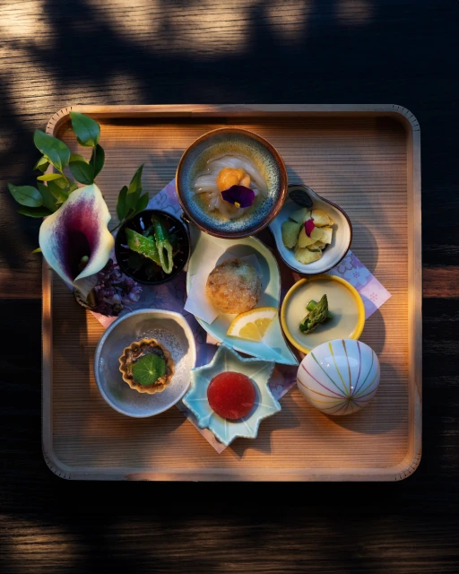 Overhead shot of an artful, Japanese-style taster plate featuring multiple small dishes, flowers, and decorative elements on a wood tray.