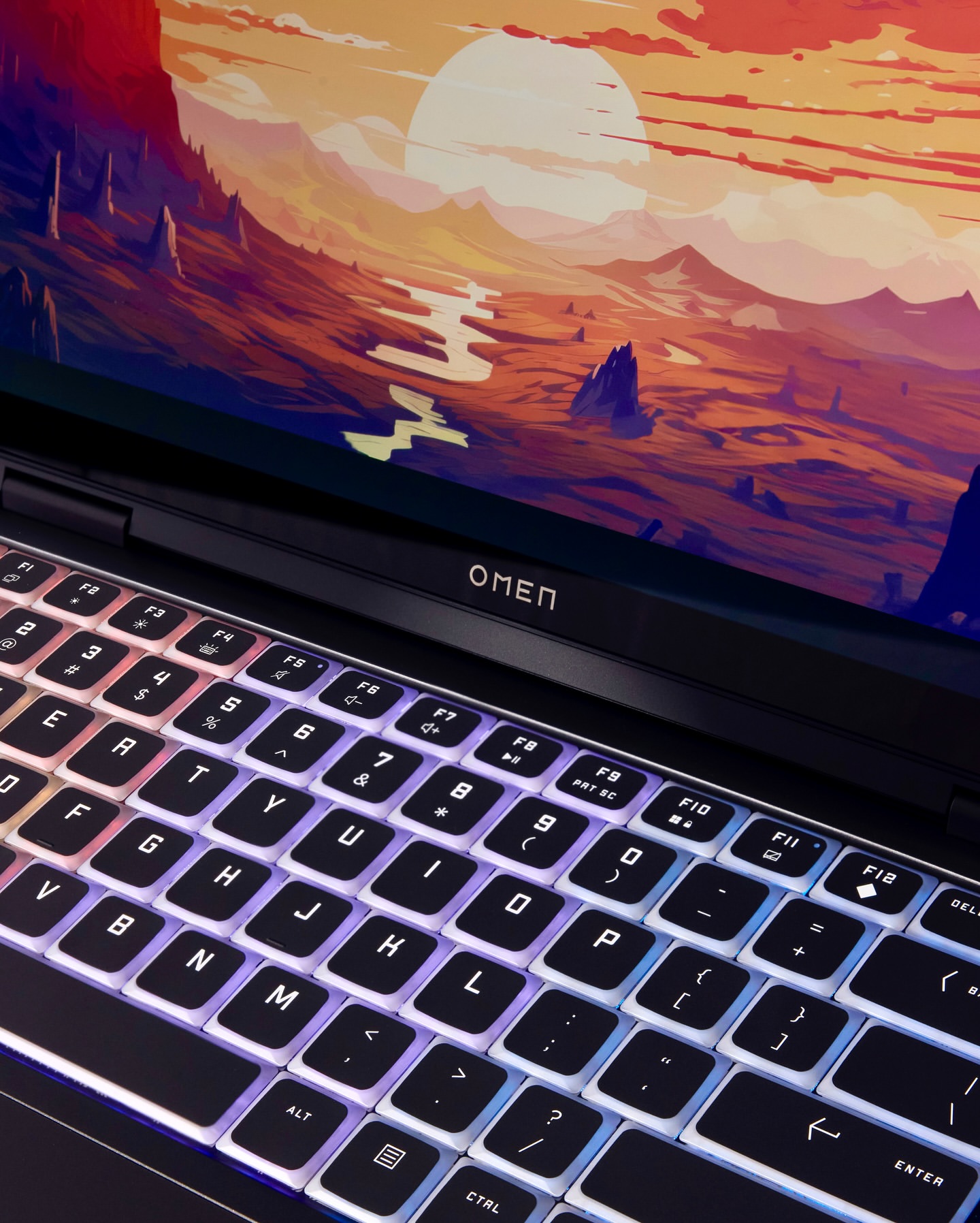 Laptop displaying a landscape art wallpaper with mountains, a river, and a large sun. Backlit keyboard of the computer is visible.