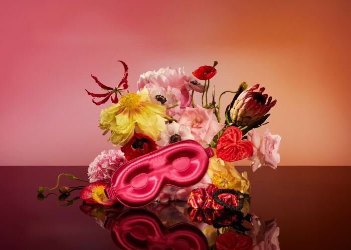 Still life of a pink satin sleep mask and a hair tie surrounded by colorful roses, poppies and exotic flowers in front of an orange background.