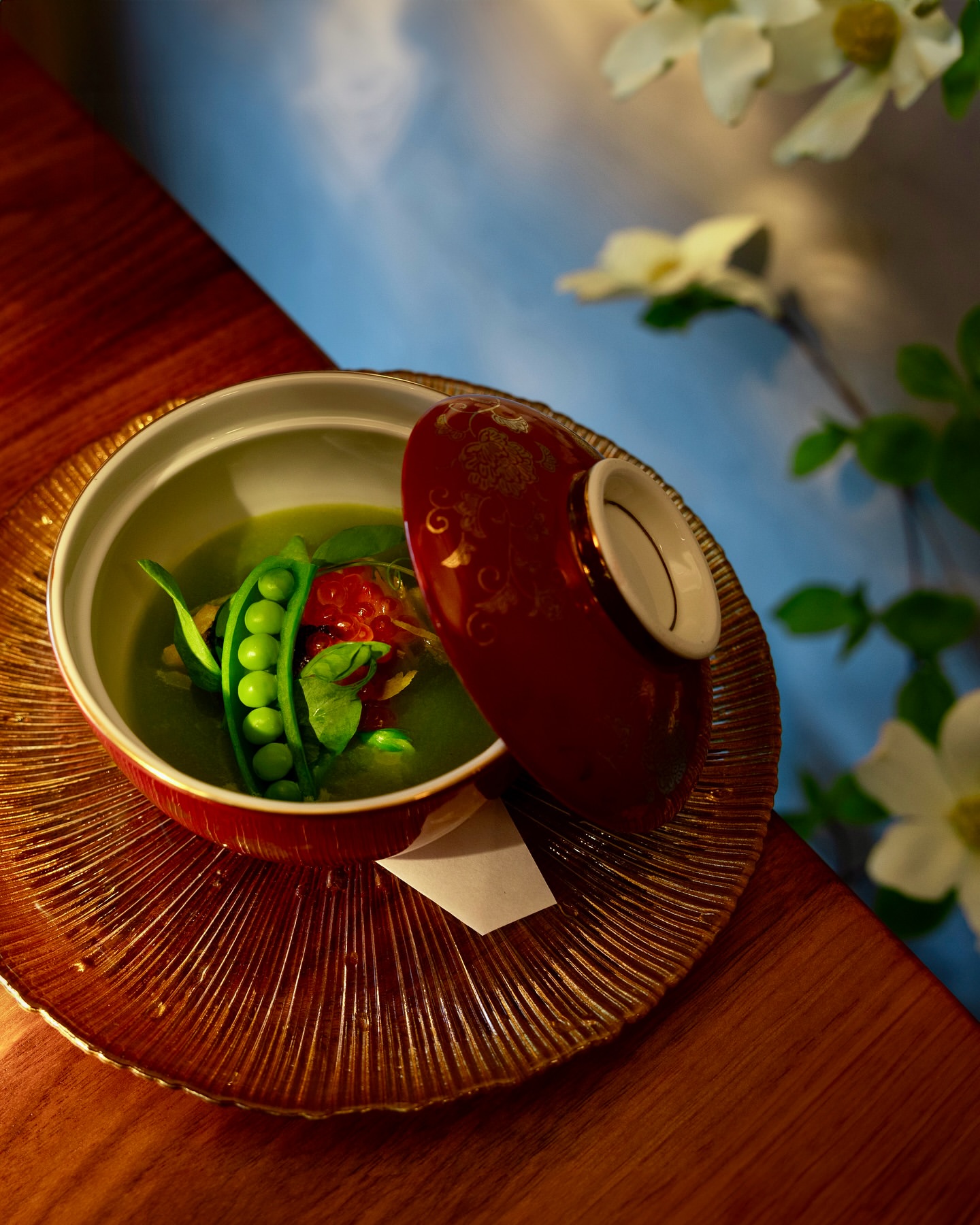 Overhead shot of a bowl of green soup with fresh peas and tomato, presented on a decorative glass plate next to white flowers.