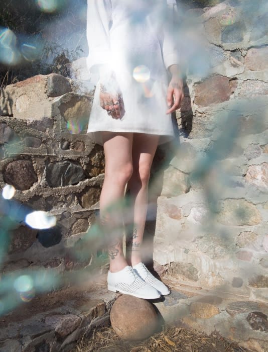 A slender adult in a white dress and perforated white shoes standing on a rock in a stone ruin area, with bright light and water effects.