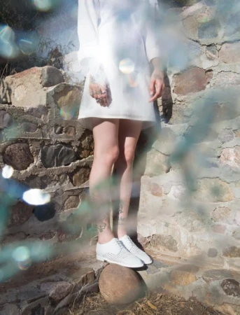 A slender adult in a white dress and perforated white shoes standing on a rock in a stone ruin area, with bright light and water effects.