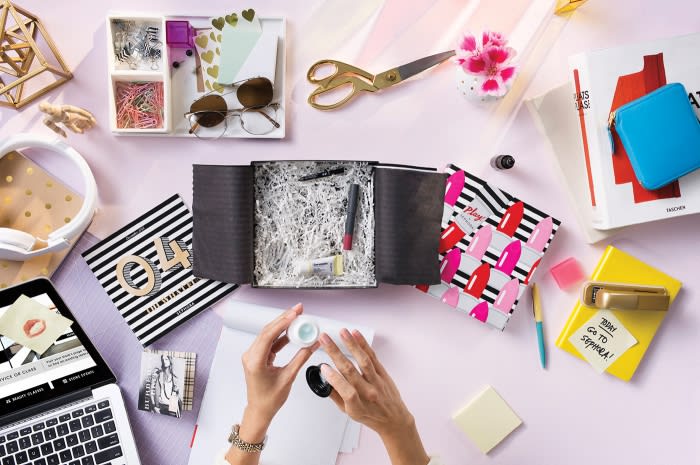 Overhead shot of a woman's hands opening a makeup subscription box with beauty products, stationery, laptop, and desk accessories.