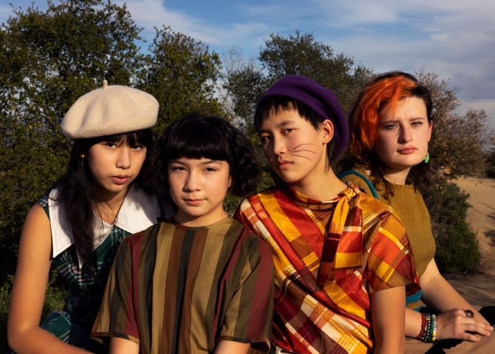 Four young women pose outdoors in vintage-inspired clothing against a backdrop of trees and a bright sky, creating a fashion-forward group portrait.
