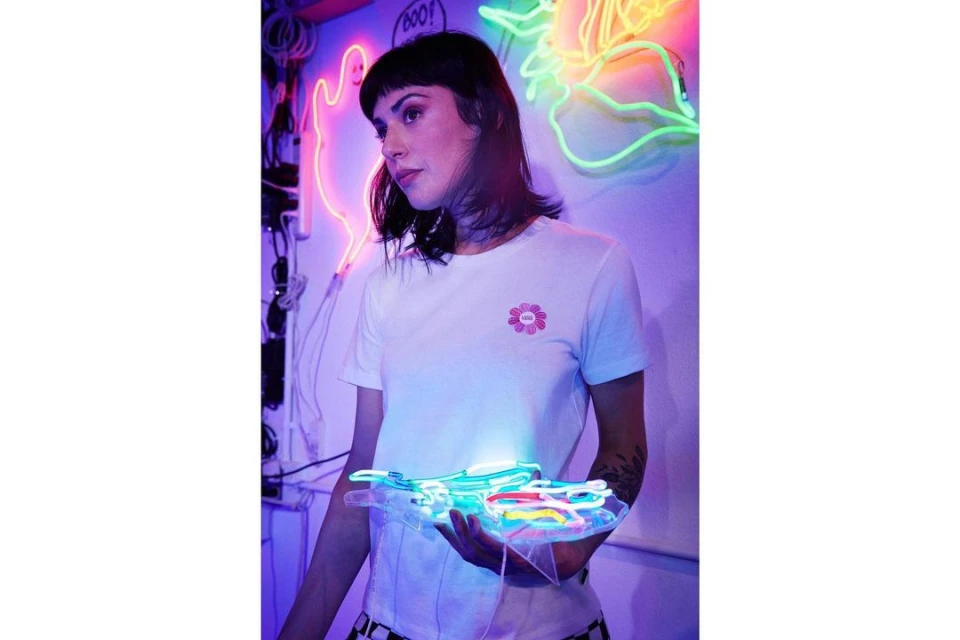 A woman with dark hair holds a neon sculpture in a room with neon lighting and visible wiring, wearing a white t-shirt.