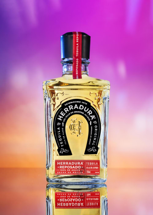 Herradura Reposado tequila bottle against a vibrant purple and pink backdrop, showcasing its golden liquor and distinctive horseshoe logo.