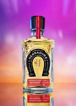 Herradura Reposado tequila bottle against a vibrant purple and pink backdrop, showcasing its golden liquor and distinctive horseshoe logo.