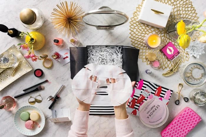 Overhead shot of person holding sheet mask amid cosmetics, jewelry, and self-care products on a marble surface, promoting skincare and beauty.