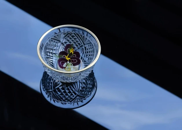 Close-up of a crystal bowl with gold trim, holding a delicate food creation decorated with edible purple and yellow flowers on a dark surface.