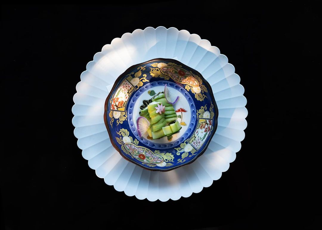 Overhead shot of sliced cucumbers and decorative flower in a floral patterned bowl, presented on an ornate white plate against black.