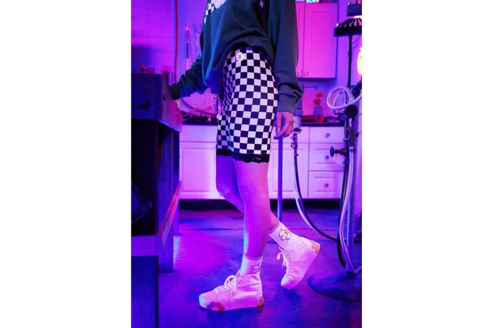 Indoor shot of an adult wearing a green sweatshirt, checkered shorts, hi-top sneakers, and socks, posed in front of a kitchen.