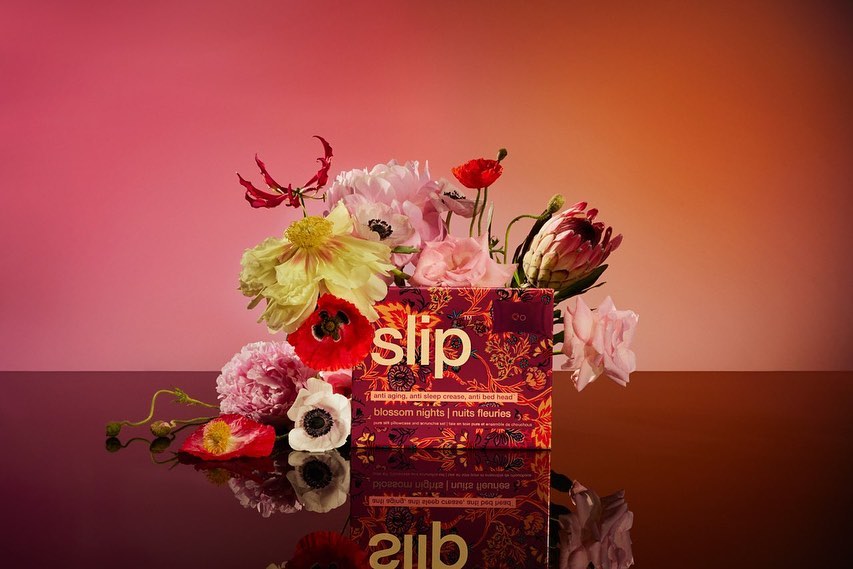 Product shot of a Slip brand box surrounded by flowers in a studio setting with pink and orange gradient lighting.
