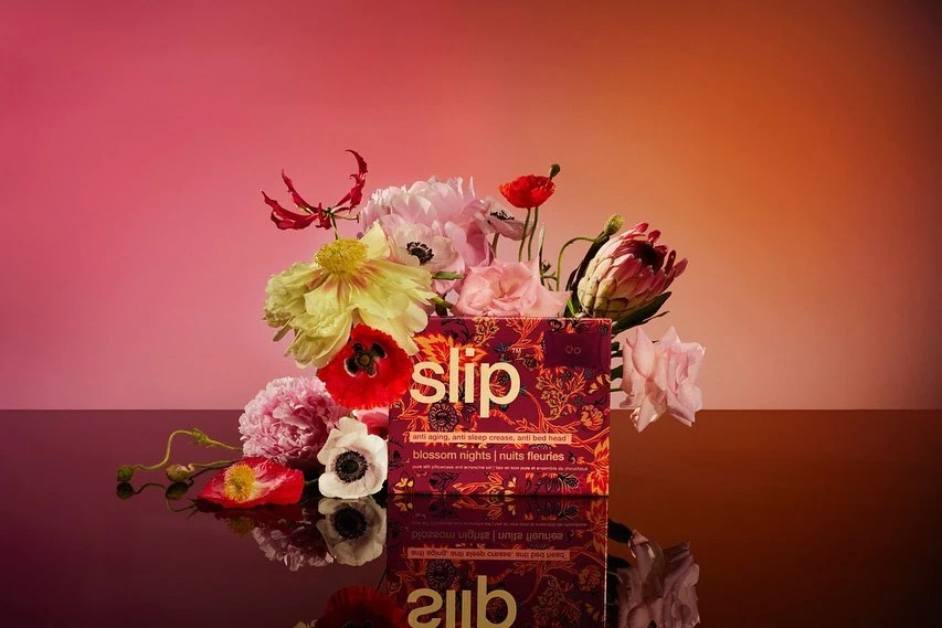 Product shot of a Slip brand box surrounded by flowers in a studio setting with pink and orange gradient lighting.