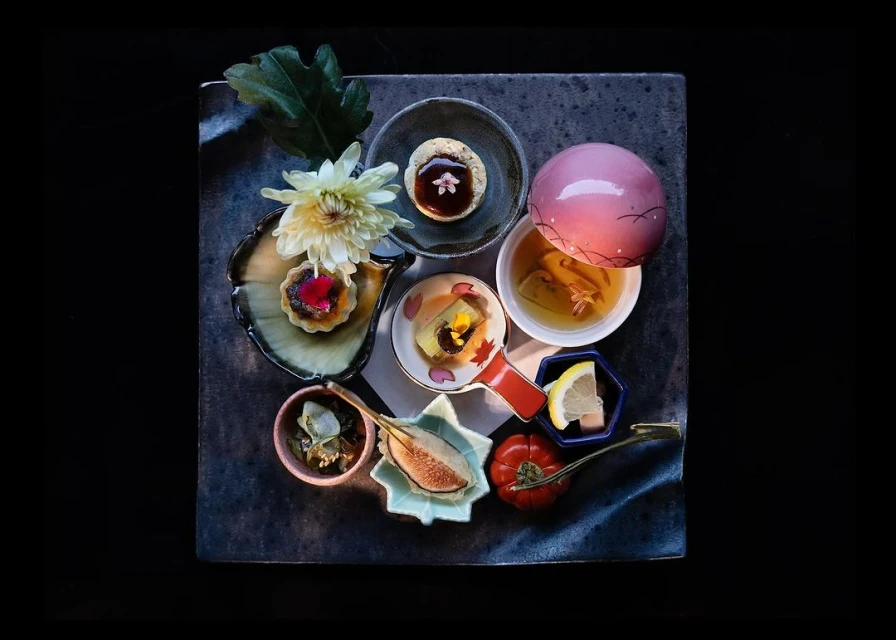 Overhead shot of vibrant, artfully arranged Japanese Kaiseki cuisine on a dark stone plate with colorful ingredients and floral garnishes.