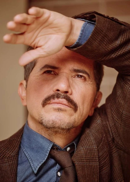 Portrait of a middle-aged man shielding his eyes with his hand, wearing a brown tweed blazer, blue denim shirt and patterned necktie.