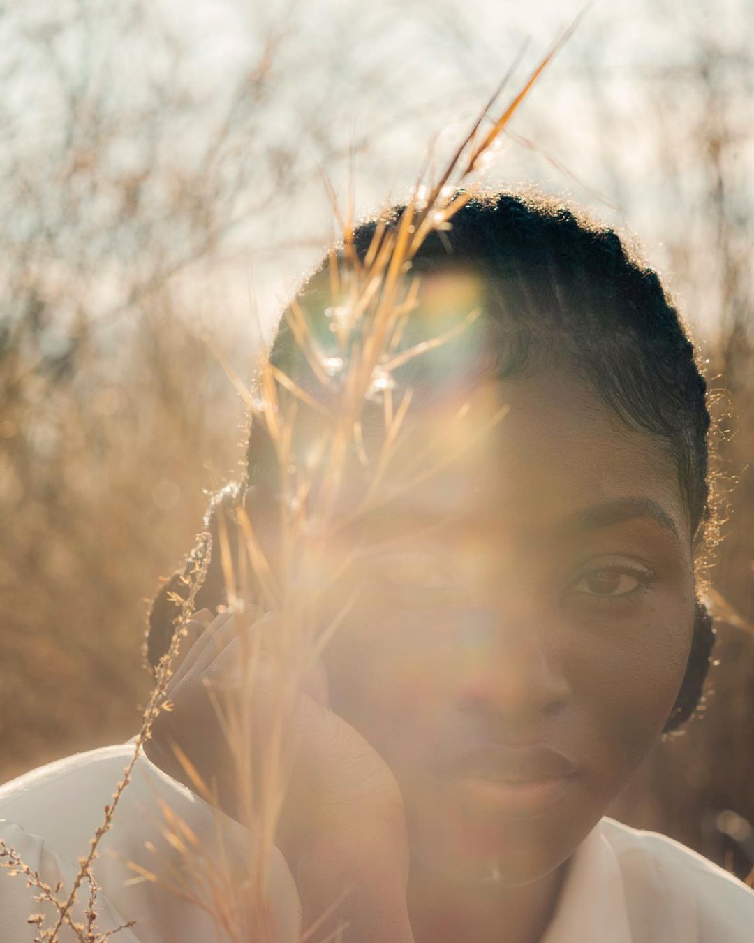 Portrait of a black woman with dark hair, posing outdoors among dry grass in warm lighting, wearing a white shirt.