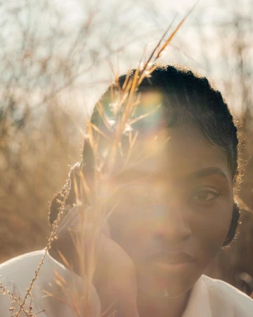 Portrait of a black woman with dark hair, posing outdoors among dry grass in warm lighting, wearing a white shirt.