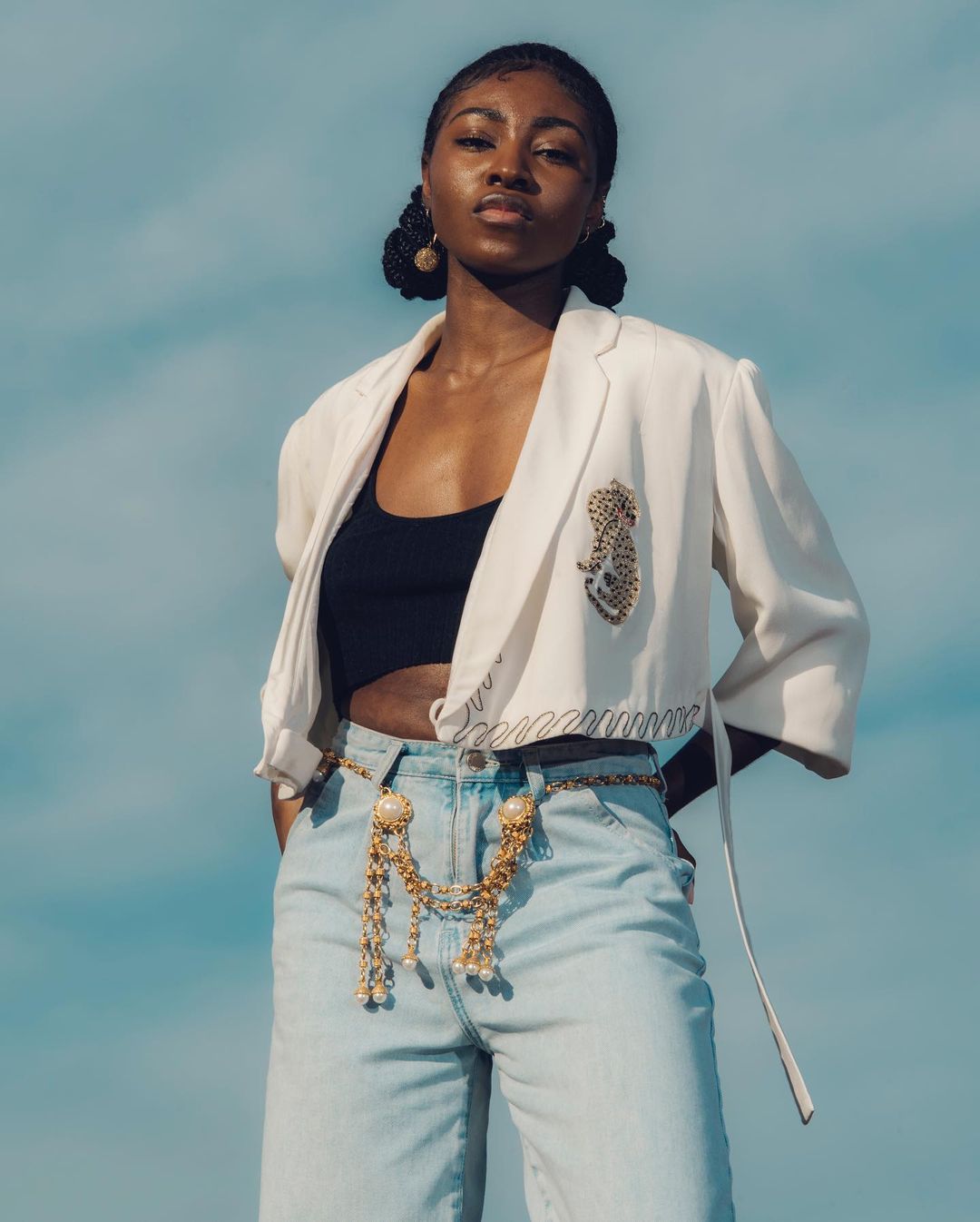 A fashionable young woman poses outdoors against a blue sky, wearing a white blazer, black top, gold belt, and blue jeans.