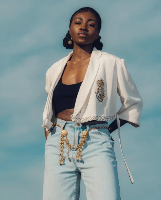 A fashionable young woman poses outdoors against a blue sky, wearing a white blazer, black top, gold belt, and blue jeans.