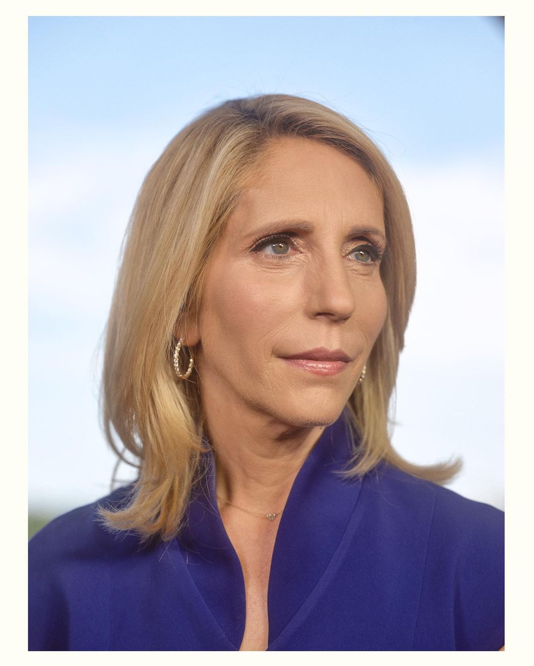 Portrait of a blond woman wearing a blue blazer against a soft blue sky background. Her hair is shoulder-length and she's wearing hoop earrings.