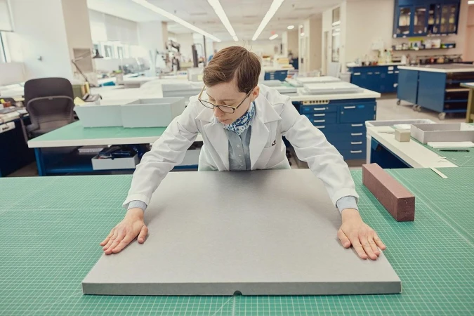 Adult woman in a lab coat working with a gray slab on a green cutting board surface, surrounded by lab equipment in a bright room.
