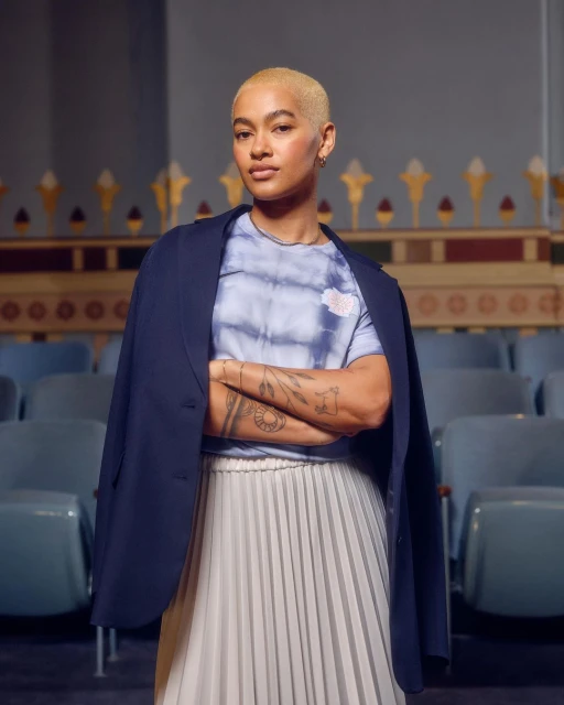 Woman with short blonde hair stands in a theatre wearing a blazer over a tie-dye t-shirt and white pleated skirt, arms crossed.