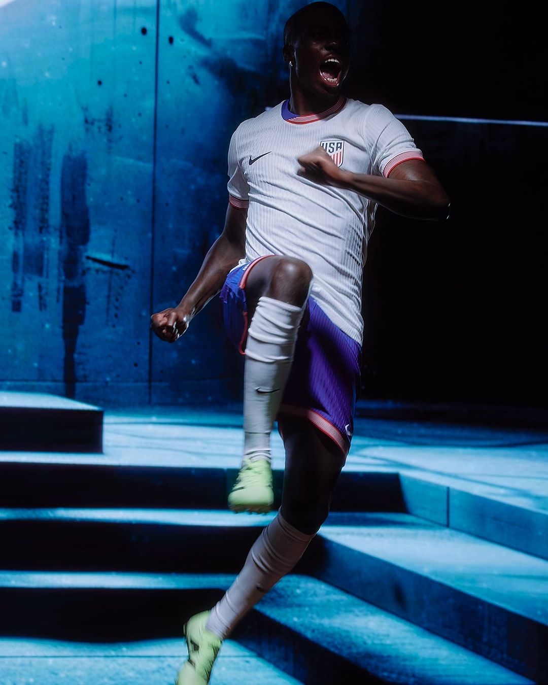 Excited soccer player in uniform celebrates a win with fist raised, standing on concrete steps in front of a textured blue wall.