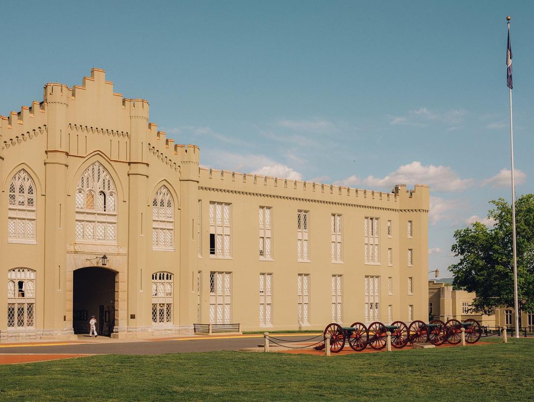 Historic military college building with turrets, arched windows, and cannons on a green lawn under a blue sky with a flag.