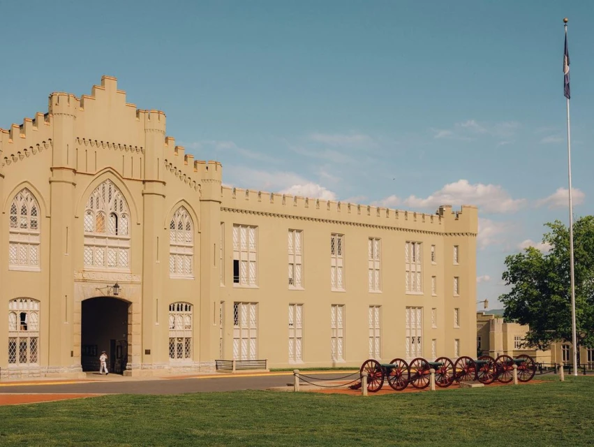 Historic military college building with turrets, arched windows, and cannons on a green lawn under a blue sky with a flag.
