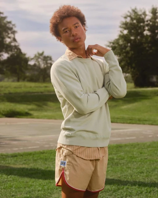 A young adult with an afro poses outdoors wearing a light green sweater, collared shirt, and red-trimmed tan shorts in a relaxed setting.
