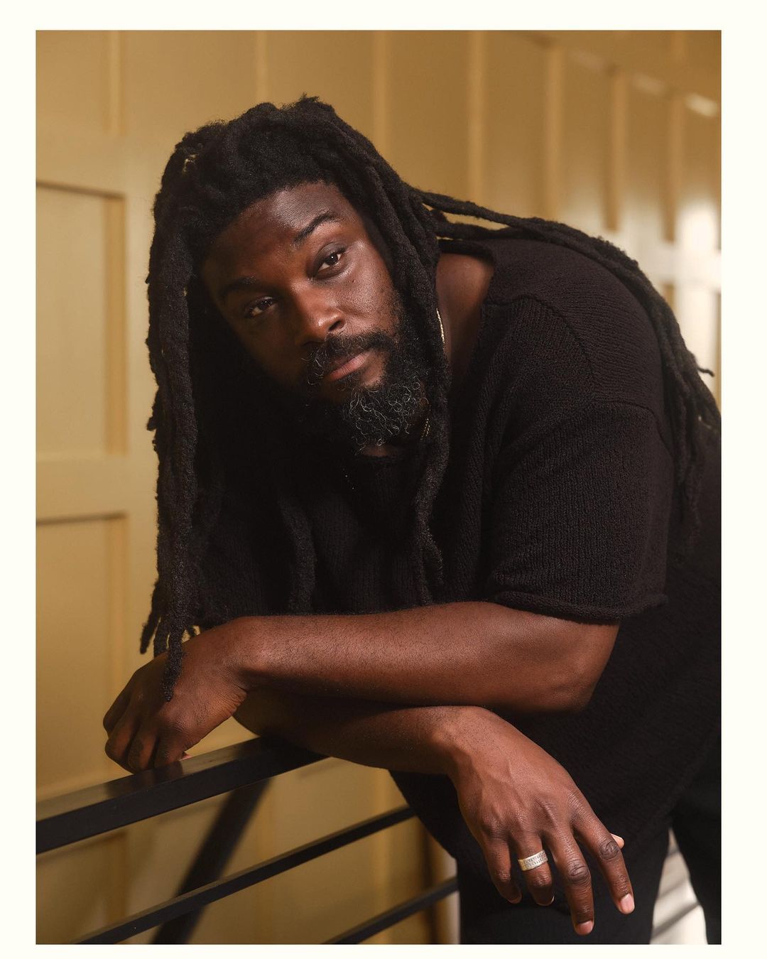 Portrait of a Black adult male with dreadlocks wearing a black shirt, resting his arm on a railing indoors against a neutral background.