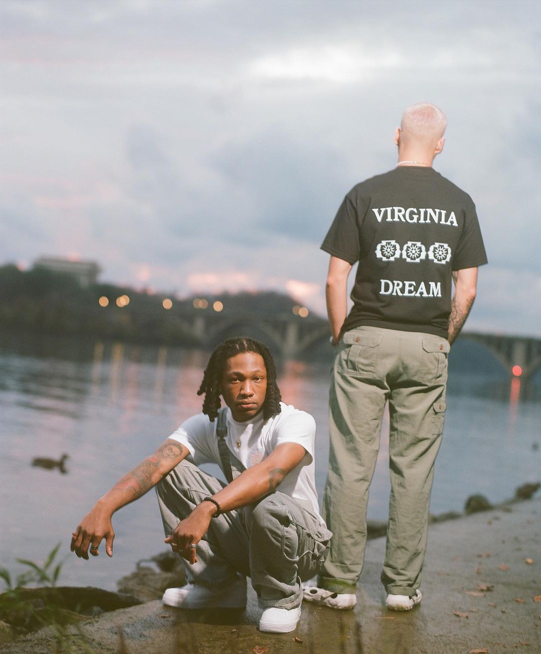 Two men pose by a river at dusk. One man with dreadlocks squats while the other stands wearing a Virginia Dream t-shirt.