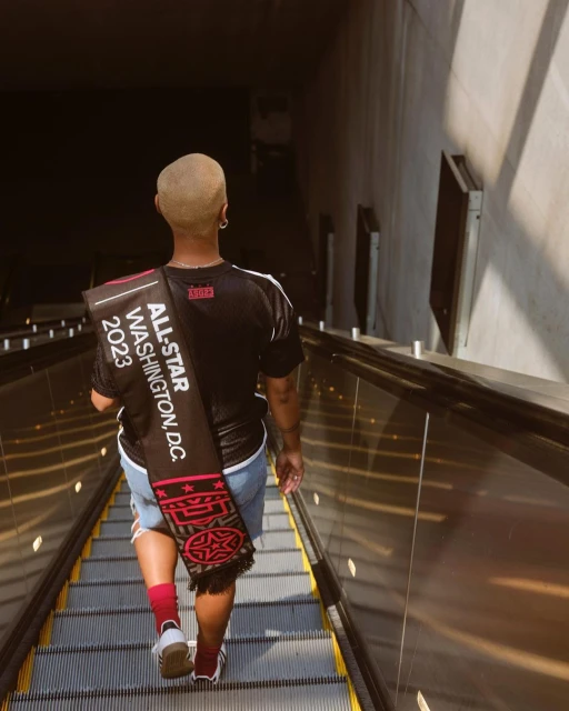 A person walks down an escalator wearing a black jersey, jean shorts, and red socks, carrying an All-Star Washington DC scarf.