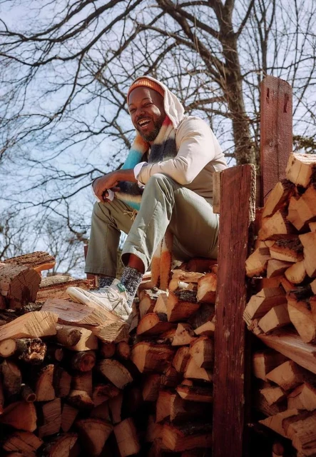 Smiling African American man sits on a stack of firewood wearing a hat and scarf in a woodland environment on a sunny day.