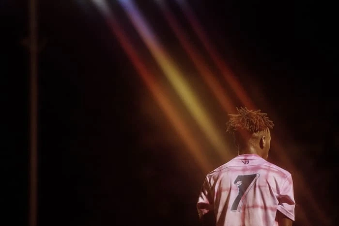 Back view of a Black adult male with orange hair in a pink jersey with the number 7, lit by colorful overhead spotlights.