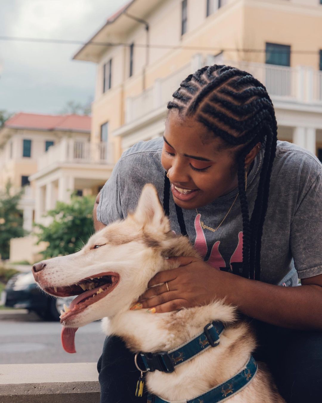 An African American woman smiles while embracing her tan and white Siberian Husky dog outdoors on a bright day in a residential area.