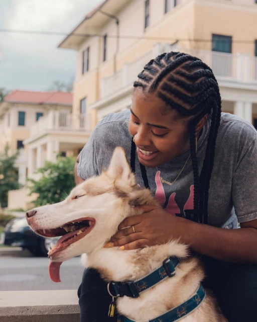 An African American woman smiles while embracing her tan and white Siberian Husky dog outdoors on a bright day in a residential area.