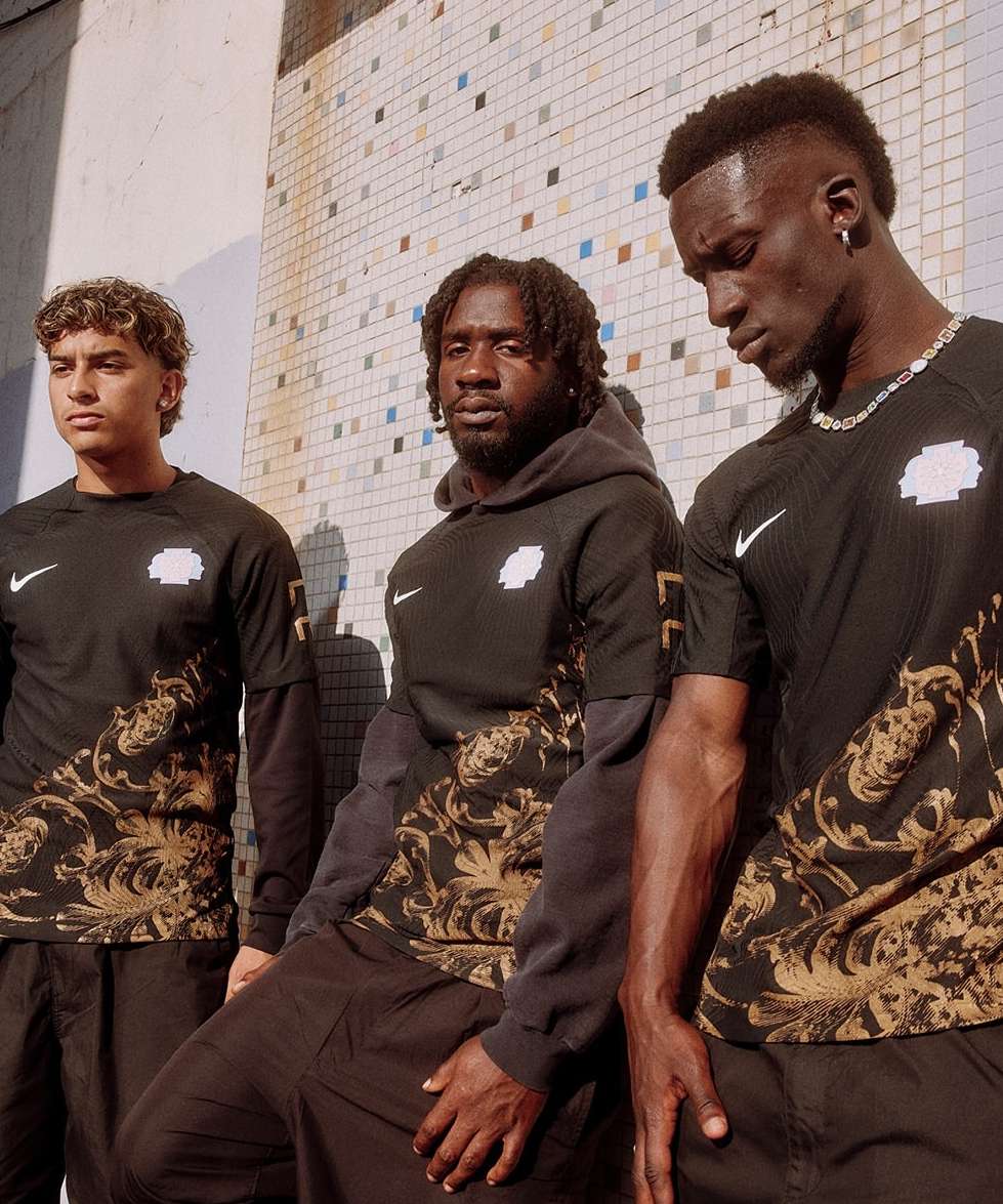 Three male models in athletic streetwear pose against a tiled wall in an urban setting, showcasing black and gold patterned jerseys.